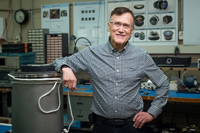 George Ricker stands in a lab, resting his elbow on a large metal cylinder.