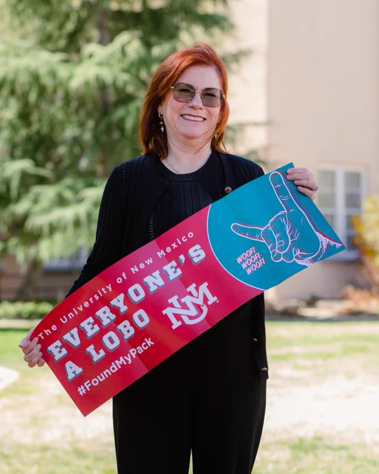 UNM President Garnett S. Stokes, dressed in black, smiles as she holds a banner reading "The University of New Mexico Everyone's a Lobo #FoundMyPack"