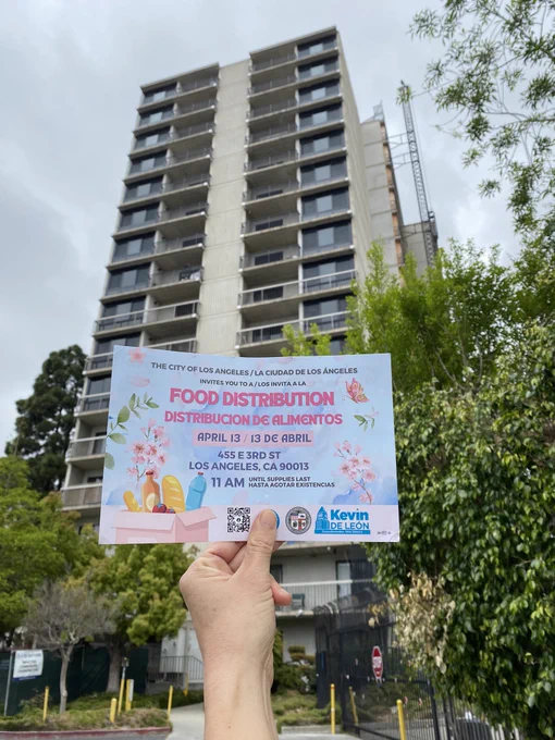Photo of a hand holding a blue flyer with pink text with a grey building in the background.

Text reads:
“THE CITY OF LOS ANGELES /LA CIUDAD DE LOS ÁNGELES

INVITES YOU TO A / LOS INVITA A LA
FOOD DISTRIBUTION DISTRIBUCION DE ALIMENTOS

APRIL 13 / 13 DE ABRIL
455 E3RD ST
LOS ANGELES, CA 90013
11 AM UNTIL SUPPLIES LASS
HASTA AGOTAR EXISTENCIAS

Kevin De Leon”