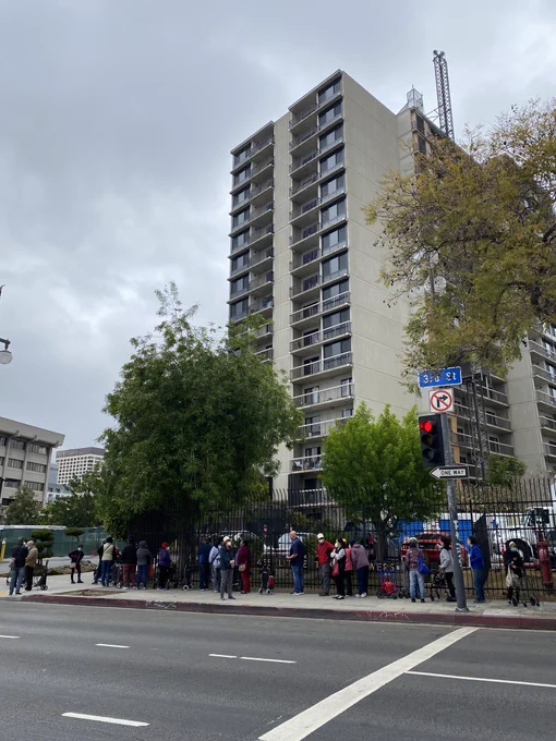 Photo of a line of little Tokyo elders standing along the sidewalk of 3rd street. In the background is a grey building.