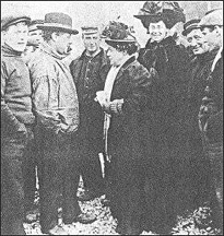 Smiling women in rather elaborate nats talking to some bemused or amused working men.