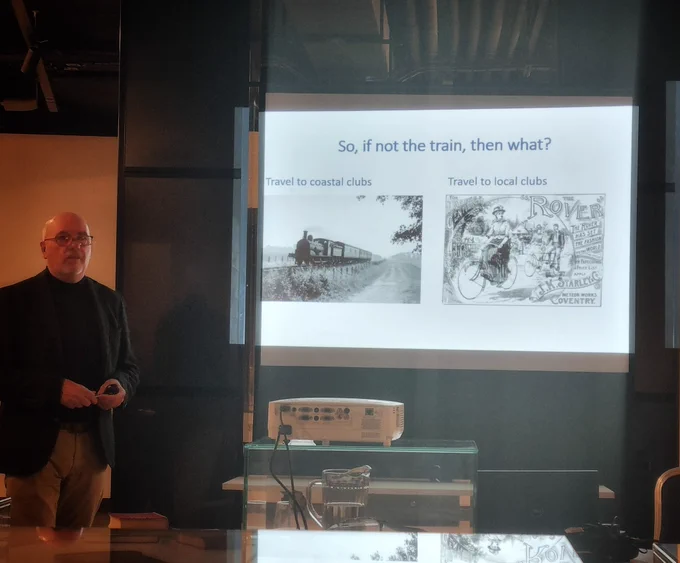 A speak is addressing the audience at a Museum lecture.  His slides how is displayed behind him.  On the screen are images of a steam train and a cartoon of a female cyclist from the Edwardian era.