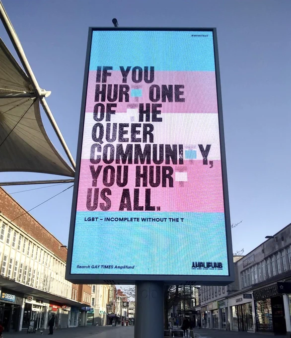 A street walkway with a sign centred and large in focus that reads:
“If you hurt one of the queer community you hurt us all.”
All of the letter ‘t’s were removed from this sentence. The sign continues:
“LGBT - incomplete without the T
Search GAY TIMES amplified”