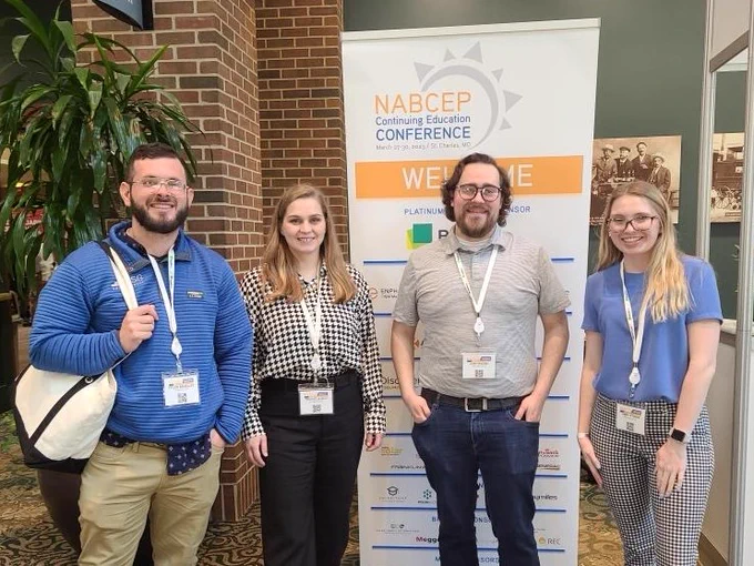 Joshua Raebel, Libby Stokes, Tyler Brady, and Michaela Bell are smiling and standing together beside a placard for the NABCEP 2023 CE Conference.