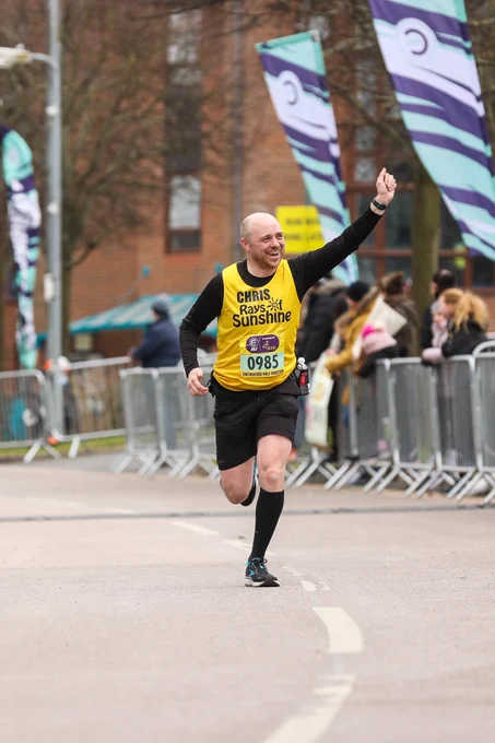 Dad wearing a yellow running vest which says Chris Rays of Sunshine, waving to the crowd while running and smiling
