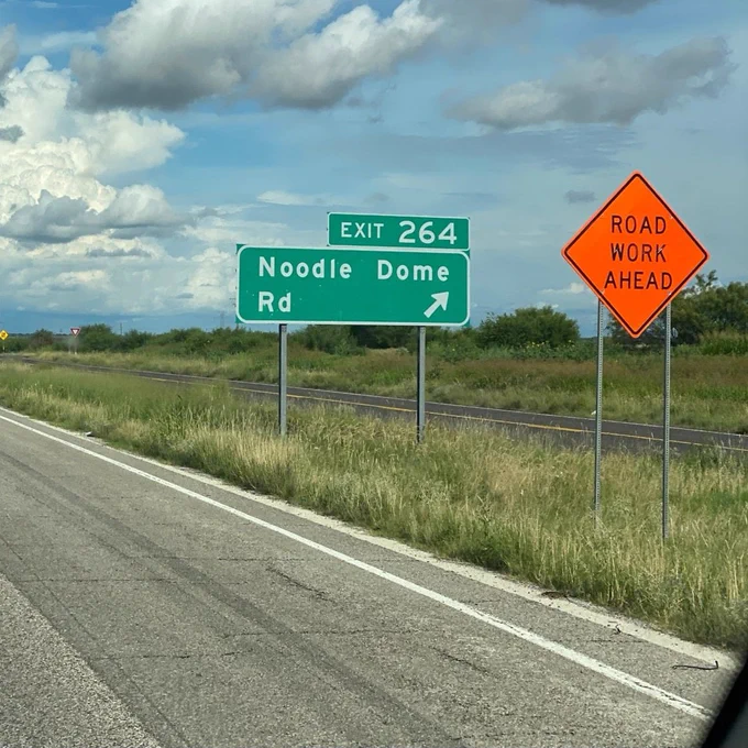 An orange roadside is on the side of the highway with the text "road work ahead" (which is quickly becoming the state flag of Texas), and a green road sign behind it says "noodle dome road, exit 264." Grass, trees, road, and a partly cloudy sky can be seen in the background 