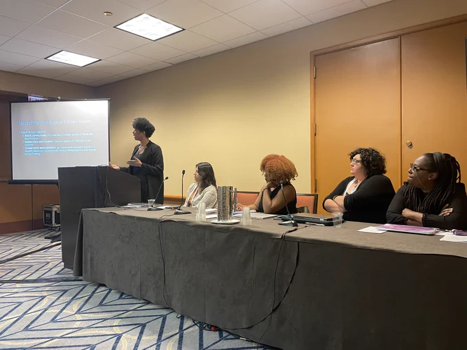 Mahasan Chaney staring at the podium with slide projected on a screen to the left and other panelists seated to the right (René Espinoza Kissell, Rachel Williams, Jackie Bristol, and Terrenda White)
