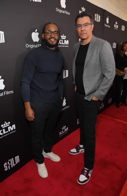 Red carpet setting. On the left is Ryan coogler dressed in a black long sleeve, pants and black framed glasses. On the right is Pete Nicks dressed in a gray suit with black pants and framed glasses.