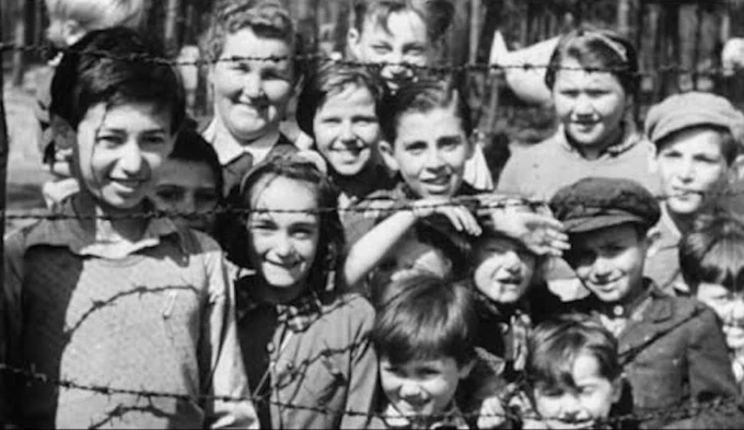 A group of children stand behind barbed wire. Second from left, standing in the back, is a woman, amidst the children.