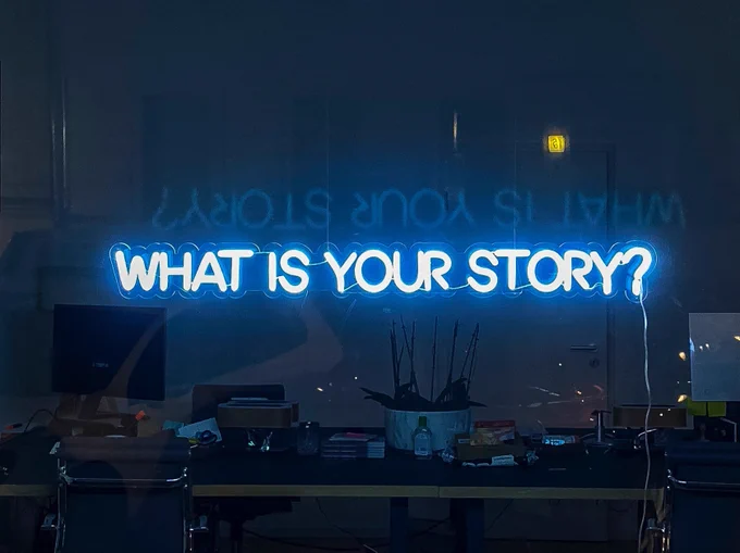 Blue neon lights in a darkened window read “What is your story?”