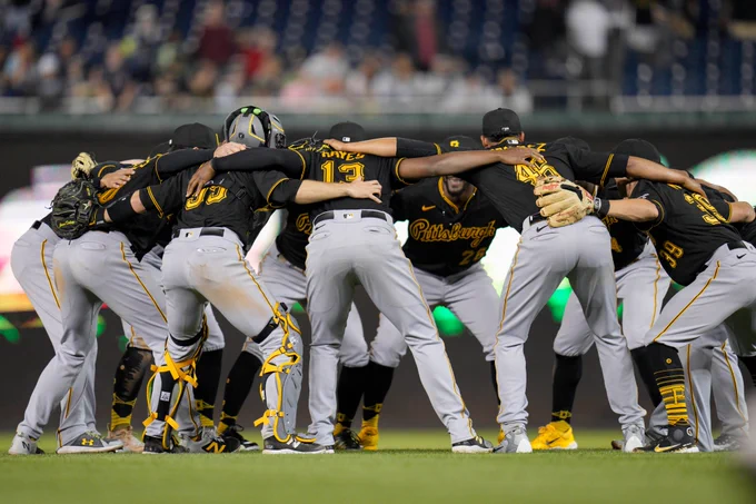 The Pirates celebrate after their 20th victory standing in a circle with their arms around each other wearing their black jerseys with gold letters and numbers with gray pants.