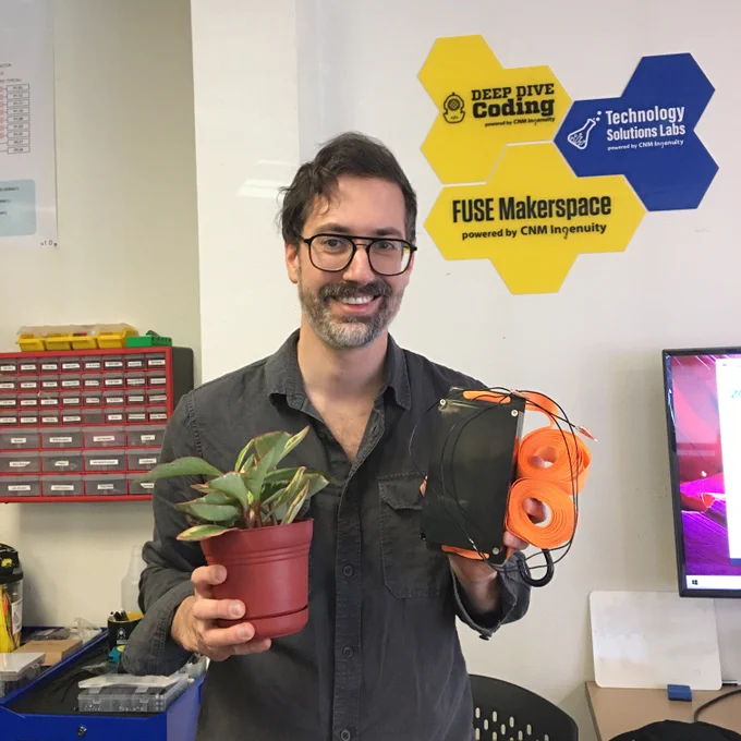 A man in a gray button-up smiles at the audience holding a few parts of his project, a small plant in one hand and a sensor box with wires in the other hand.