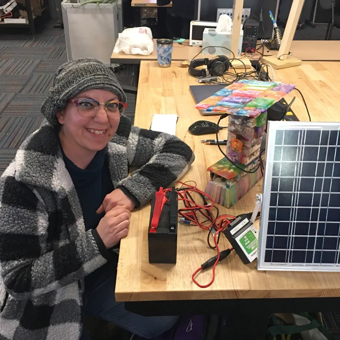 A woman wearing a beanie hat and checkered Sherpa jacket smiles at the camera while seated next to part of her project, a solar panel connected to a battery.