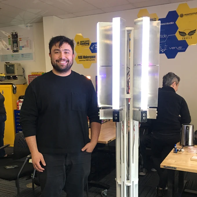 A man in a black long-sleeve shirt and black jeans stands proudly next to his IOT project, a stand with vertical lights and sensors.