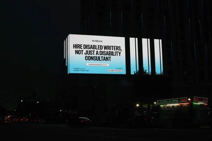 A large billboard, lit up at night over a street. It shows bold black text over a white and blue gradient background that reads: Hey Hollywood, Hire Disabled Writers, Not Just A Disability Consultant