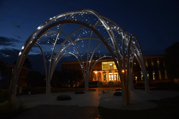 The arches of the memorial are connected with tiny lights that shine against the night sky like stars. The entrance of Kennedy is lit in the background.