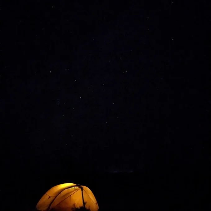 An image of a line tent, illuminated from the inside, set against a jet black sky with stars twinkling above. The tent appears green-orange in the light...