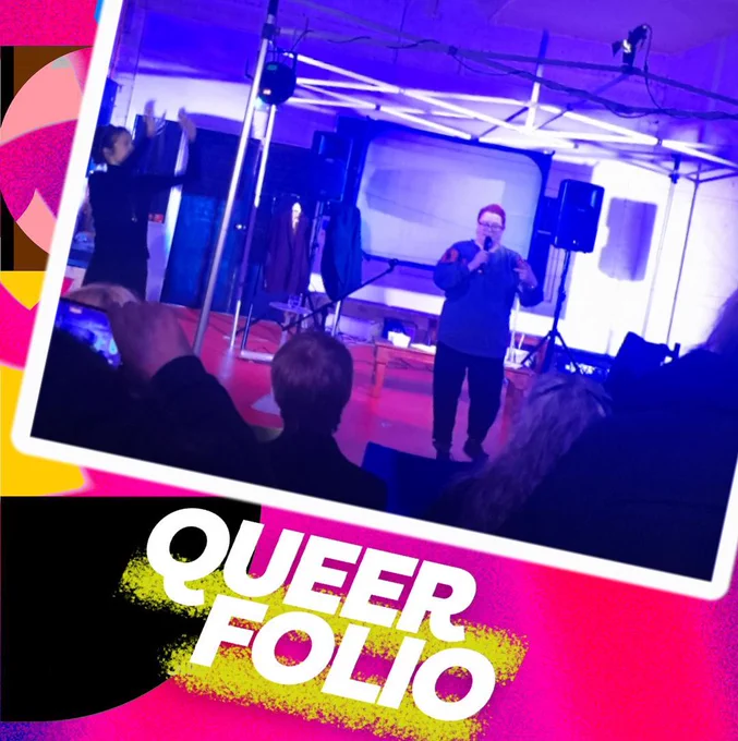 Inside a studio, a person with pink hair holds a microphone and speaks to an audience. To their left is a person who signs.