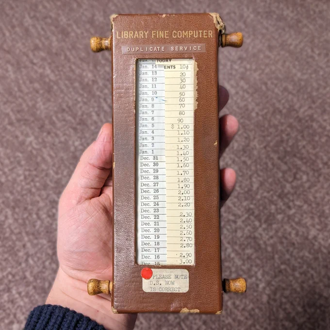 Close up of hand holding worn case with four wood knobs and  a long list of dates with another list beside it of costs that increase by ten cents per day