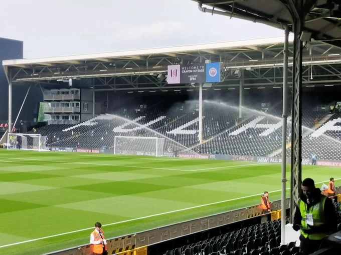 Photo of the Craven cottage pitch with the sprinklers on and the Hammersmith End in the background with the screen showing the logos of Fulham and Manchester City 