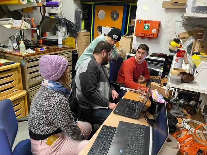 Das Foto zeigt drei Menschen vor einem Arbeitstisch mit aufgeklappten Laptops und Tastaturen sitzend. Dahinter lehnt sich eine weitere Person an eine Sessellehne. Die Leute unterhalten sich. Auf dem Arbeitstisch befinden sich zwei Filamentspulen und zahlreiche weitere Gegenstände. Über dem Arbeitstisch befindet sich ein Wandregal mit Kartons und zahlreichen weiteren Gegenständen. Im Hintergrund stehen Regalsysteme mit vielen Laden und darauf ein 3D-Drucker. Weiters ist ein oranger Erste-Hilfe-Kasten an der Wand und eine hell-orange Türe mit runden spiegelnden Flächen und eine Ampel zu sehen.