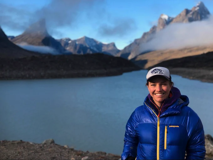 Photo of a smiling Sarah Crump in ball cap and puffy coat with a stunning arctic landscape behind her of lake, jagged peaks, and low clouds.