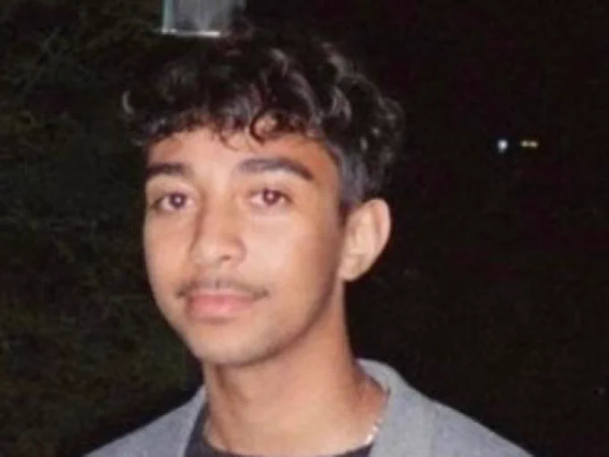 A person with short hair looks at the camera. The background is black. 