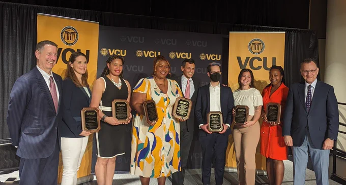 A group of winners of the VCU Presidential Awards for Community Multicultural Enrichment pose with their awards on a stage in front of a VCU background.