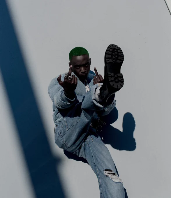 Ashton Sanders wearing an all denim look with ripped jeans, cuffed pants, a white t shirt, and black boots. The denim jacket has a quilted puffer quality to it. His accessories include a lion and one tiger shaped ring on his right hand, and a simple band and circle shaped ring on the left. Ashton has dyed green afro hair in a buzz style. He is kicking his right leg up casting a shadow on the wall behind him of his leg and leaning back while flipping off the camera with both hands. Next to him is the shape of a shadow in a somewhat wide streak on his right side