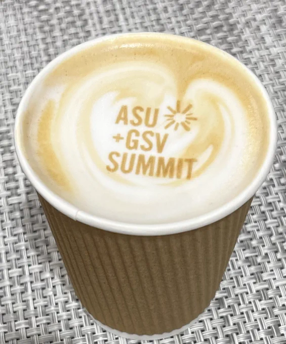 A cup of coffee in a paper cup displays the ASU   GSV Summit logo in edible printing on milk foam.