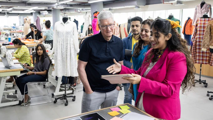 Tim stands with three students at the Indian School of Design & Innovation. Everyone looks at an iPad as one student speaks while pointing to the screen. In the background, several students work at their desks, surrounded by mannequins displaying a variety of fashion designs.