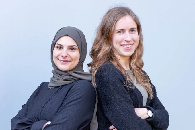 Portrait of Seinab Noura (left) and Sophie Farkas (right), standing back to back with their arms crossed and smiling into the camera