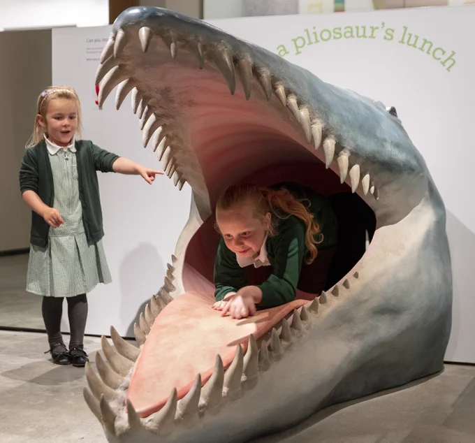 Model of pliosaur with mouth wide open showing teeth. Inside the mouth is a young school girl, another is standing beside it pointing. Courtesy of Dorset Museum. 