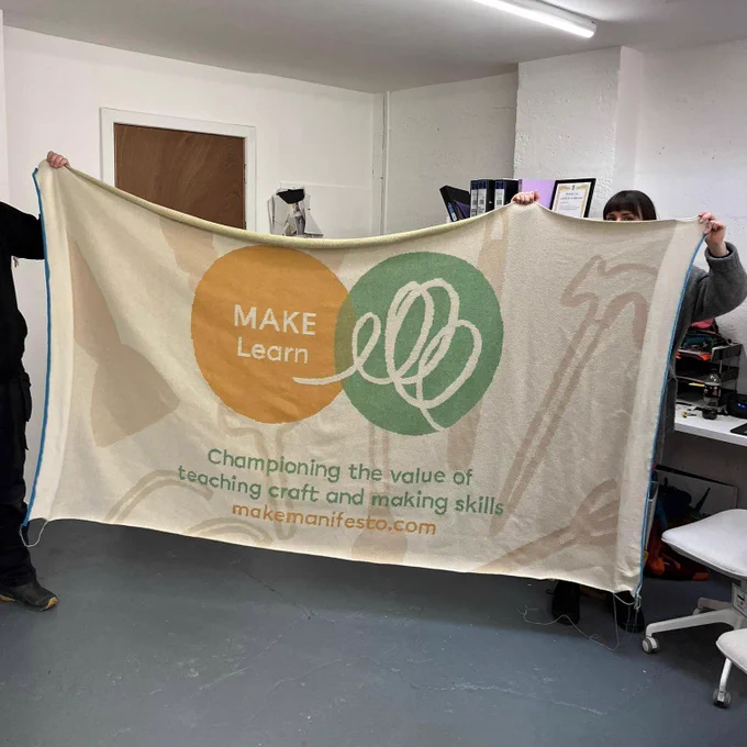Two figures hold up a very large knitted banner that reads: "MAKE Learn: Championing the value of teaching craft and making skills. makemanifesto.com"