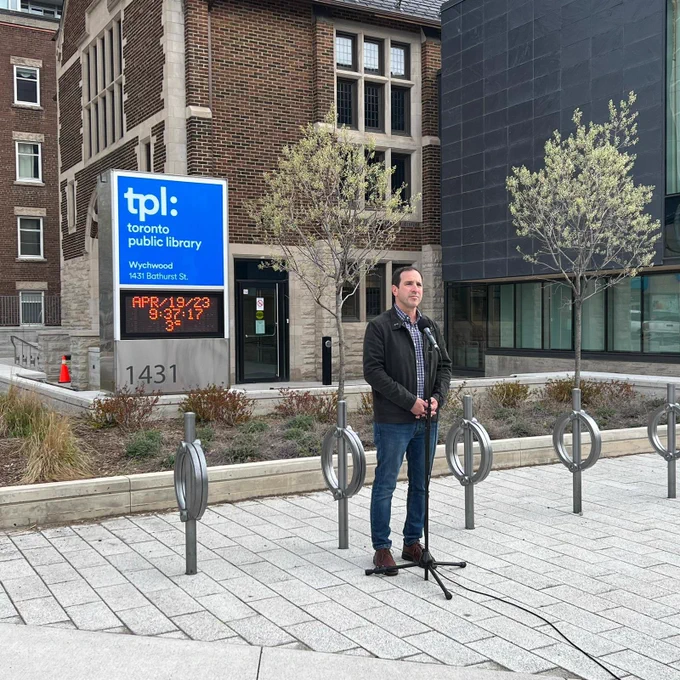 A white man with a grey jacket, checkered shirt and blue jeans at a microphone standing in front of a library building