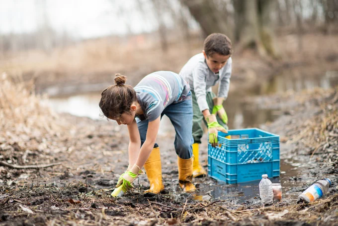 Two children spend time on a mucky spring day as they clean up garbage and recyclables from the mud and puddles around them. They are wearing rubber boots and gloves as they work together to put items in a blue crate.