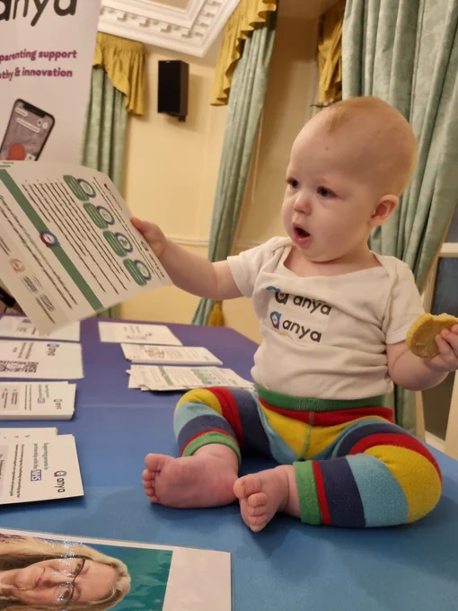 A baby in an Anya branded vest and stripey leggings hands out leaflets at the Anya baby & breastfeeding app exhibition stand.