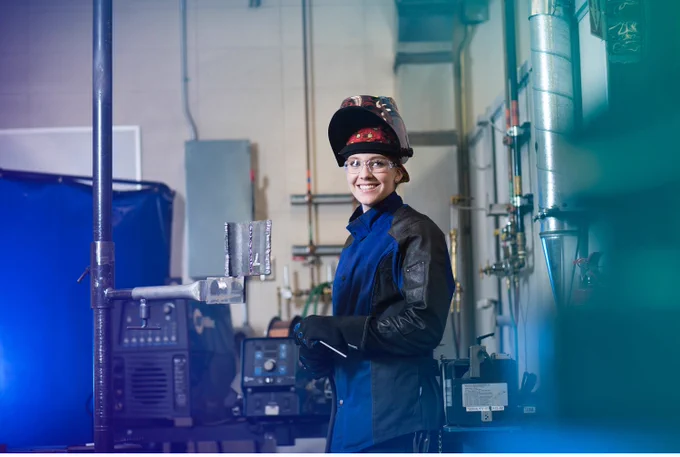 Woman in welding gear stands beside a welder. 