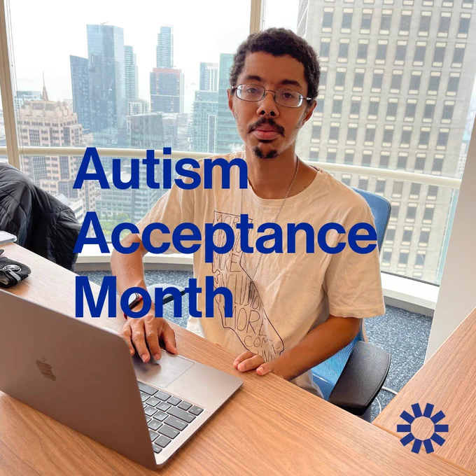 Isaac, a light brown man with a beard and glasses, sits at a desk. The city of San Francisco can be seen through windows in the background. The text on the image reads "Autism Acceptance Month." The blue Kelsey logo is visible in the lower right-hand corner.