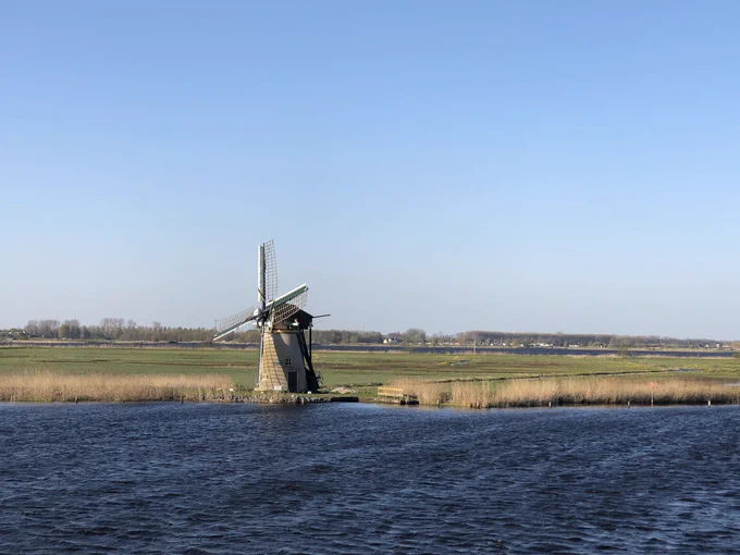 Photograph of a small windmill on the edge of a river, with very flat fields behind it and barely any gap to the edge of the water. The sky is clear and pale blue, and the water is dark blue with lots of ripples and small waves.