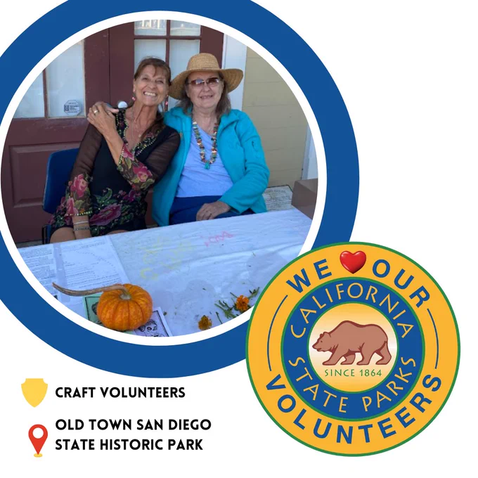 Circle image of two Craft Volunteers at Old Town San Diego State Historic Park. They are sitting behind a table that has a pumpkin on it. Below that to the right is the round California State Parks Logo with "We Love Our Volunteers" around the outside. Word Love is replaced with a heard symbol. There is a shield icon at the bottom left. Next to it reads "CRAFT VOLUNTEERS".
Below that is a location icon. Next to it reads "OLD TOWN SAN DIEGO STATE HISTORIC PARK".