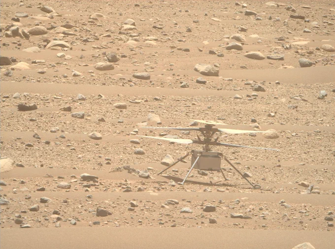 Photo from NASA’s Mars Perseverance rover shows the Ingenuity Mars Helicopter on the Red Planet. The small, dual-rotor helicopter rests on four thin legs and is seen in the lower right of the photo, on a dusty, rock-strewn surface. In overhead sunlight, the helicopter casts a shadow directly below. Its horizon-facing terrain camera can be seen at the center-bottom of the fuselage. A thin layer of dust can be seen on its rotors and solar panel.