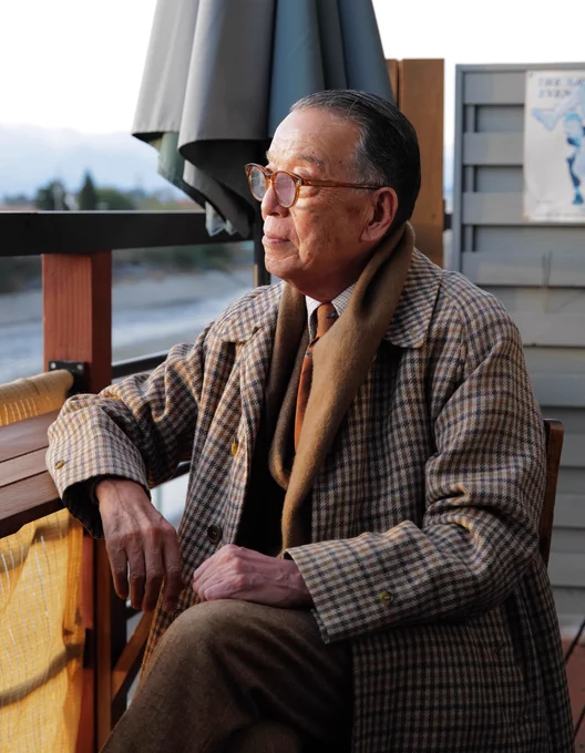 Yukio Akamine wears a brown checked tweed, raglan sleeve topcoat with slash pockets. He pairs it with a brown scarf, white tattersall dress shirt, brown flannel suit, and orange wool tie