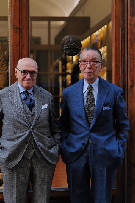 Yukio Akamine stands with bespoke tailor Antonio Liverano. Liverano is wearing a three-piece grey pupptooth, single-breasted suit with a light blue checked shirt and solid blue silk tie. Akamine wears a blue flannel, single-breasted suit with white-and-blue striped shirt and black paisley tie
