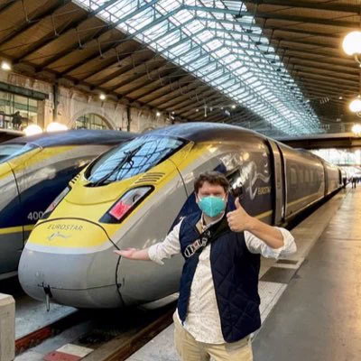 The author standing on a platform in front of a Eurostar high-speed rail train in Paris’ Gare du Nord in France before a trip through the Chunnel to London, UK, at speeds as high as 180 mph. Author has a thumb up, and is holding his other hand showing the train behind. Sunlight streams in from the station’s characteristic iron framed sky lights above in the grand vaulted ceiling.