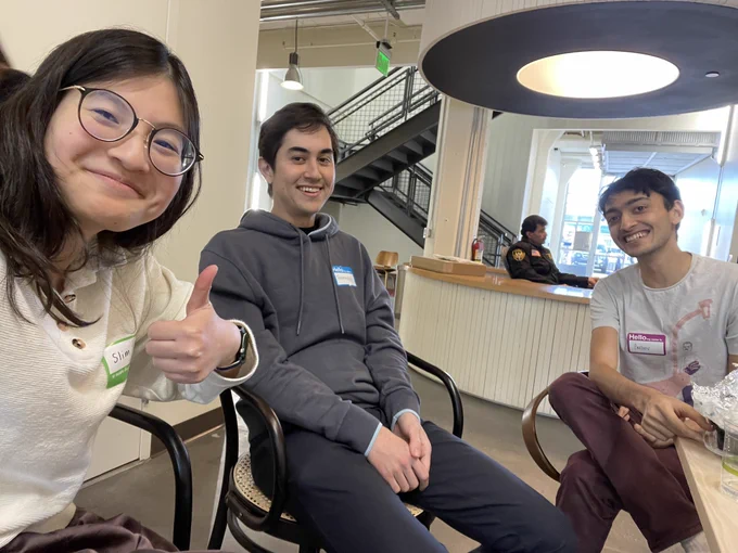 Three people sitting in designer chairs smiling bracingly: a woman with an energetic thumbs-up, a man wearing a gray hoodie with tense posture, and a joyous young man grinning beatifically (the exception to the "bracing" modifier above)