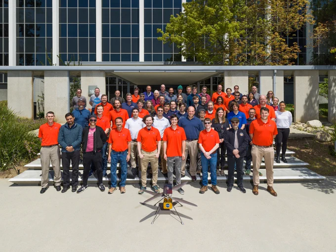 The Ingenuity Mars Helicopter team gathers at NASA's Jet Propulsion Laboratory. There are approximately 50 people standing in seven rows. Many of them are wearing red-orange team polos. A model of the Mars helicopter sits on the ground in front of them - its gold body emblazoned with the NASA logo and its rotors casting a shadow on the ground. Green bushes and trees surround the steps, and the black and white grid of building windows comprise the background.