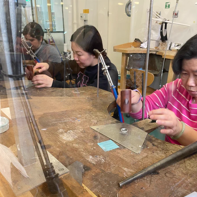 three women sit at wooden jewellery work benches. Two women are using a leather mallet to hammer rings.