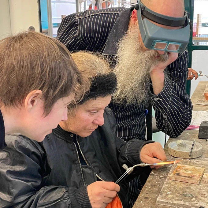 three people huddle around a workbench. The person in the centre is soldering a ring while the others look on.