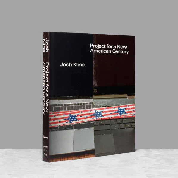The exhibition catalogue "Josh Kline: Project for a New American Century" stands against a gray background. The front of the catalogue depicts two laptops held together haphazardly by tape printed with a pattern of an American flag.
